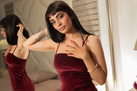 Elegant woman in a burgundy velvet dress posing in a bright, modern bedroom setting.