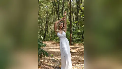 Elegant woman in a flowing white maxi dress posing gracefully in a sunlit forest setting.