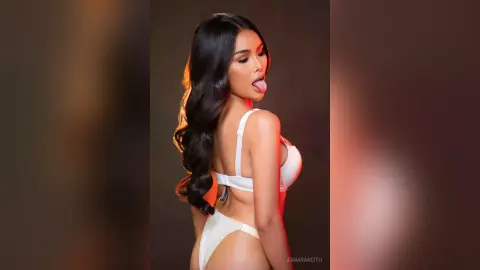 Glamorous woman in white bikini posing with long wavy hair against a warm studio background.