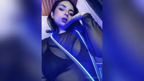 Young woman in a black sheer mesh bodysuit posing under moody blue lighting.