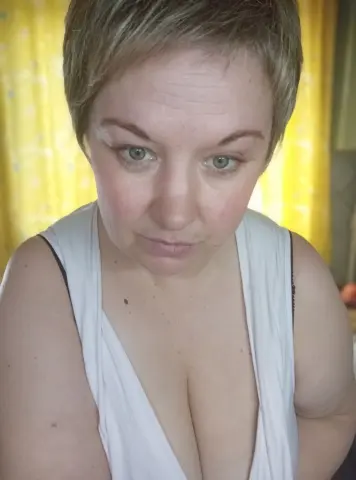 Juli_Bond looks at the camera wearing a white tank top with a deep v-neck, showing her cleavage and light brown hair.