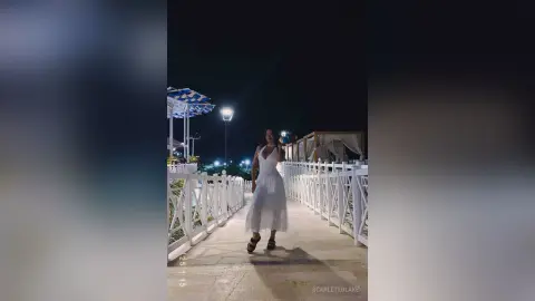Elegant woman in a white summer dress standing on a white balcony at night under soft lighting.