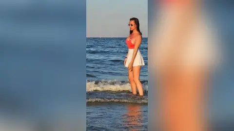 Young woman in a coral tank top and white skirt standing by the ocean waves at sunset.