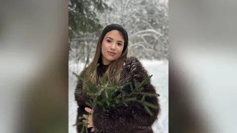Smiling woman in a cozy dark faux fur coat and black beanie holding evergreen branches in a snowy winter forest.
