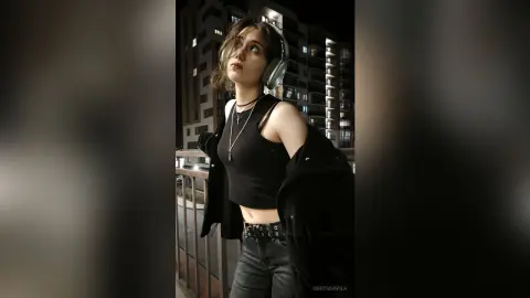 Young woman in a black crop top and headphones listening to music on a city balcony at night.