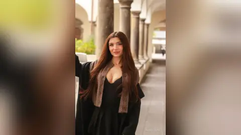 Smiling young woman in a black dress and brown cardigan standing in a sunlit classical stone courtyard.