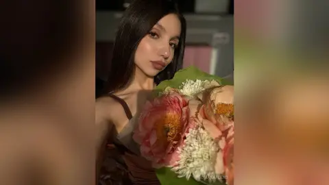 Young woman with long dark hair holding a large pink peony bouquet in soft, warm indoor lighting.