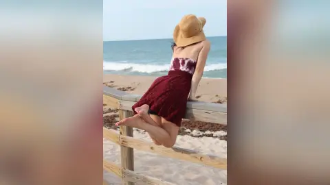 Woman in a burgundy sundress and straw hat sitting on a beach fence overlooking the ocean waves.