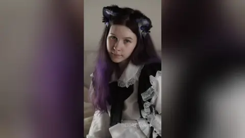 Young woman with purple hair wearing a black and white gothic lolita dress in a soft indoor setting.