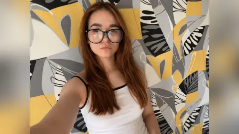 JaneBarbrick is a redhead wearing glasses and a white tank top, posing in front of a patterned yellow and grey background.