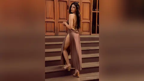 Elegant woman in a brown backless maxi dress with a high slit posing on wooden stairs in a classic setting.