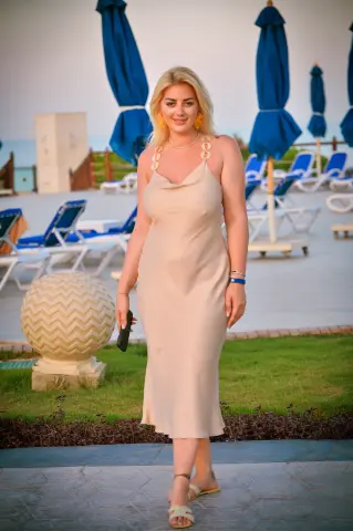 Blonde woman in a beige slip dress standing on a sunny resort patio near blue beach umbrellas.