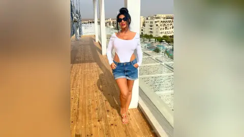 MissyJolie stands on a wooden balcony wearing a white long-sleeve bodysuit and denim shorts with sunglasses.