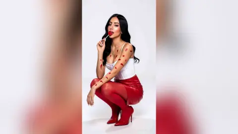 Glamorous woman in a white bodysuit and red leather skirt holding a lollipop against a white background.