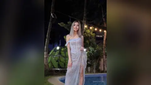 Elegant woman in a white sheer evening gown posing in a tropical garden at night.
