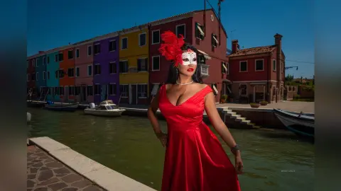 MissyJolie wears a red satin dress and a white mask while standing next to a canal in a colorful village.