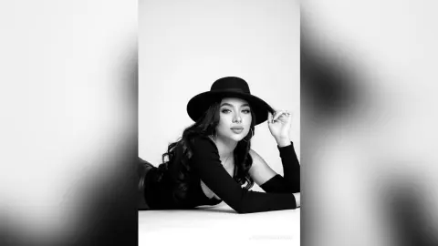 Elegant woman in a black long-sleeve top and wide-brimmed hat posing in a minimalist black and white studio setting.