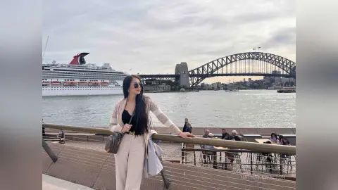 Woman in a beige cardigan and black top posing by the Sydney Harbour Bridge on a cloudy day.