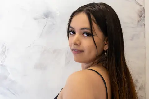 Young woman with long dark hair wearing a black tank top against a textured white wall background.