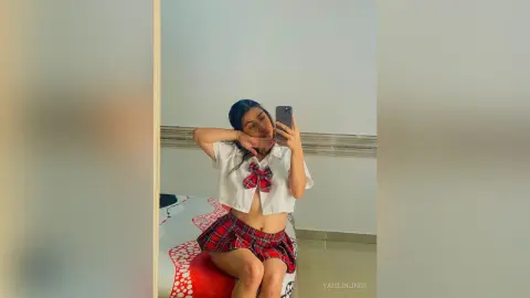 Young woman in a white crop top and red plaid skirt taking a mirror selfie in a bright room.