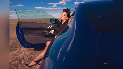 Stylish woman in a black blazer sitting in a blue car doorway on a sunny day in a rural landscape.