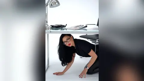 AnneThompson wearing glasses and a black t-shirt, leaning forward over a white desk with her hands resting on the surface.