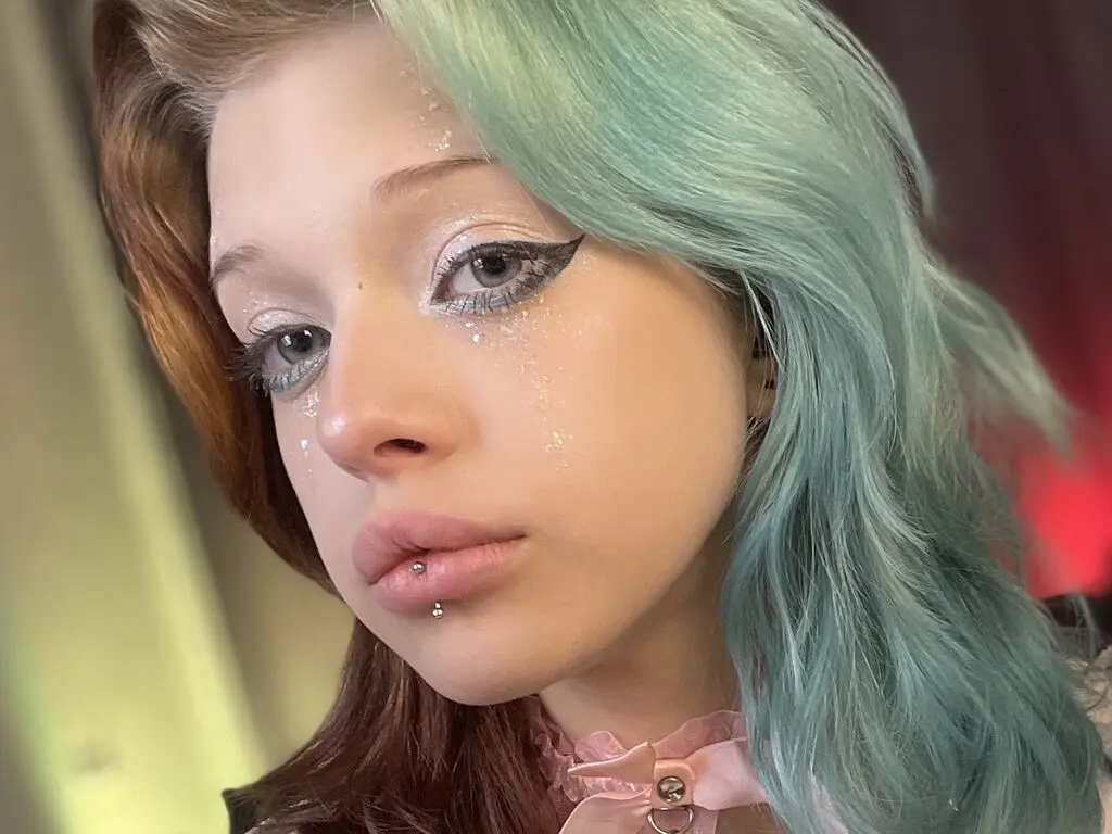 Young woman with teal and brown hair, glitter makeup, and lip piercing looking melancholic in soft lighting.