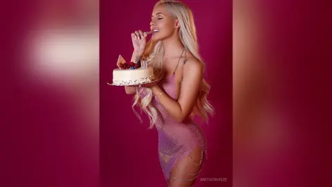 Anita poses in a pink mesh dress while holding a small cake near her mouth.
