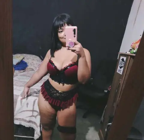 Woman in burgundy lace lingerie set posing for a mirror selfie in a dimly lit bedroom.