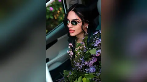 Stylish woman with dark hair and sunglasses holding a lilac bouquet inside a car during golden hour.