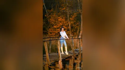 Smiling woman in white long-sleeve shirt and blue jeans standing on a wooden bridge in an autumn forest.