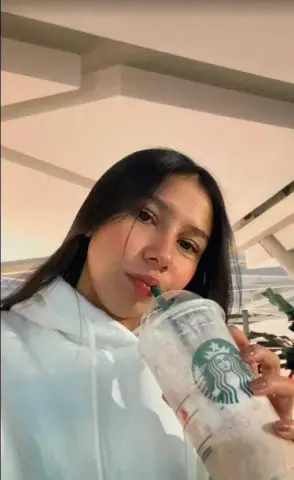 Young woman in a white hoodie enjoying an iced Starbucks drink outdoors in bright sunlight.