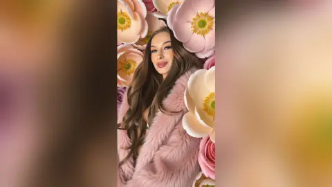 Glamorous woman in a pink faux fur coat surrounded by large soft pink flowers in a dreamy floral setting.