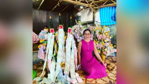 Smiling woman in a magenta dress sitting next to decorative money trees at a festive celebration.