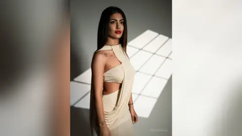 Elegant woman in a cream sleeveless cutout dress posing in a minimalist studio with dramatic sunlight shadows.