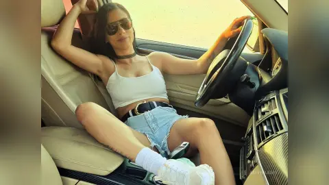 Stylish woman in white crop top and denim shorts relaxing in a car driver's seat on a sunny day.