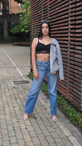 Young woman in black lace bralette, denim jeans, and grey blazer posing outdoors against a wooden slat wall.