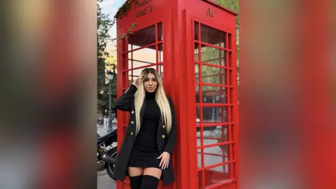 Stylish woman in a black dress and long coat posing by a classic red British telephone booth.