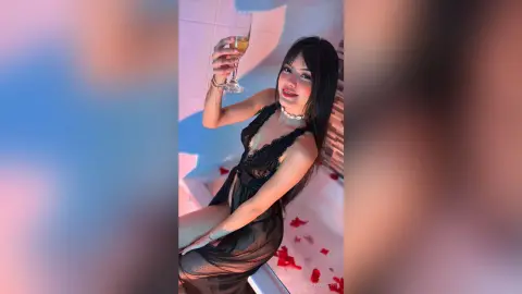 Smiling woman in a black lace dress holding a champagne glass in a moody, dimly lit room.