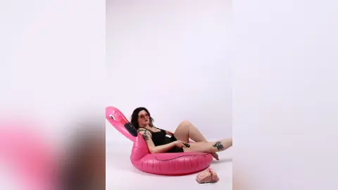 KiraKovall reclines in a pink flamingo floaty wearing a black tank top, showing off her tattoos and dark hair against a white background.