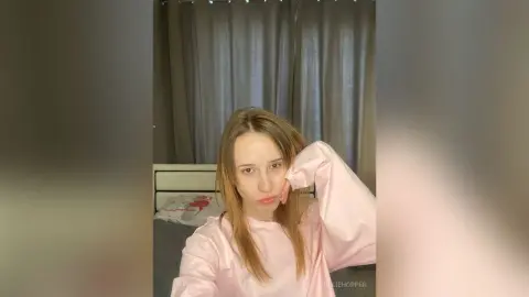 Young woman in a pink long-sleeve sweatshirt relaxing in a cozy bedroom setting.