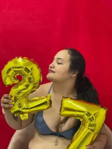 Cheerful woman in a blue bikini holding large gold number 2 balloons against a vibrant red background.