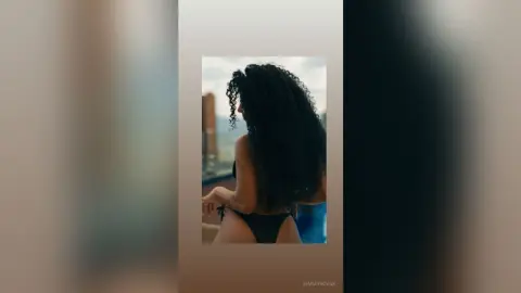 Curvy woman with long curly hair in a black bikini relaxing on a balcony with a city view.