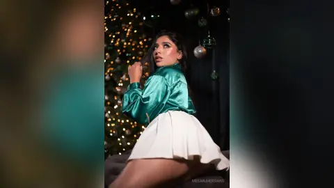 Stylish woman in a green satin blouse and white skirt posing in front of a festive Christmas tree background.