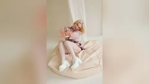 LynGoodhart sits on a beige beanbag wearing a pink dress and white patterned tights.