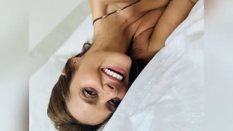 Smiling young woman in casual loungewear relaxing on white bedding in a bright, cozy bedroom setting.