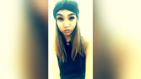 Young woman with long hair wearing a black tank top and beanie in a soft, minimalist indoor setting.