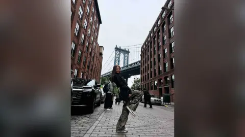 EvelynMills stands on a cobblestone street wearing a black long-sleeve top and patterned trousers with the Manhattan Bridge in the background.