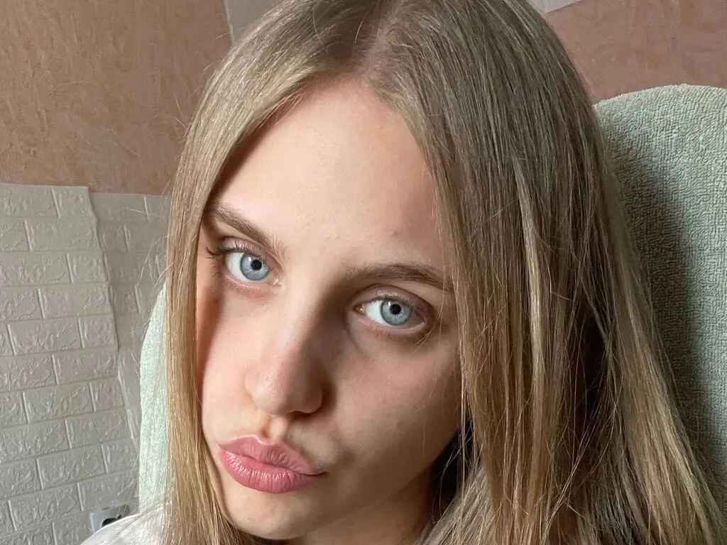 Close-up portrait of a young woman with long blonde hair and blue eyes making a playful pouty face indoors.
