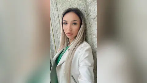 Young woman with long blonde hair wearing a white jacket and green top against a textured stone wall.